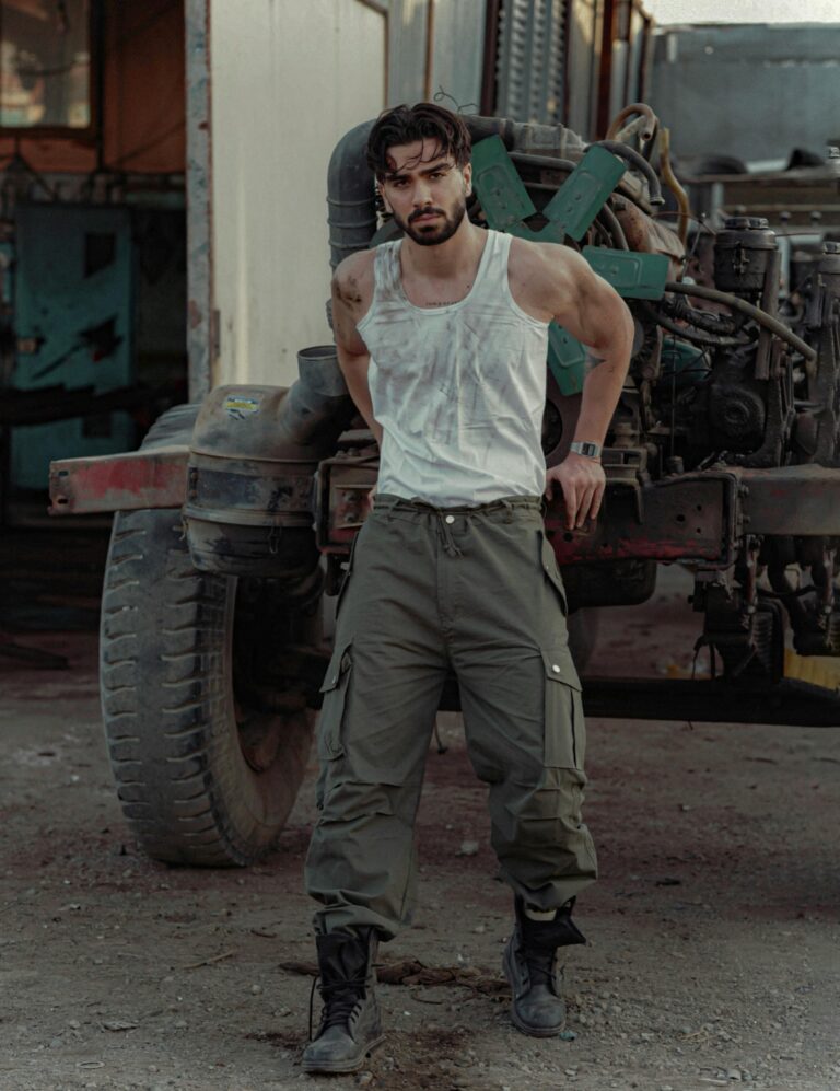 Muscular man with beard and mustache standing confidently in a garage workshop beside a rusty vehicle.