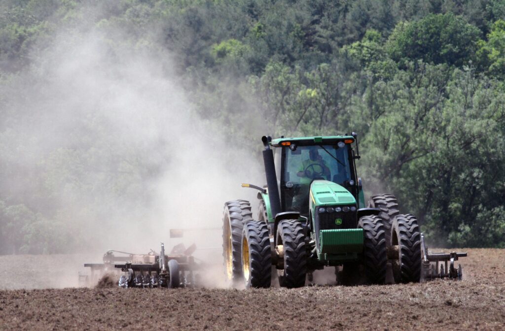 A green tractor plowing a field, creating clouds of dust in an agricultural setting.