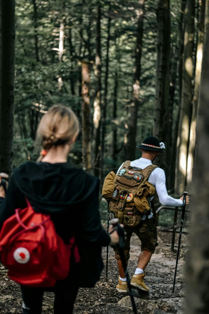 A couple hikes with backpacks and poles, exploring a dense forest on a sunny day.